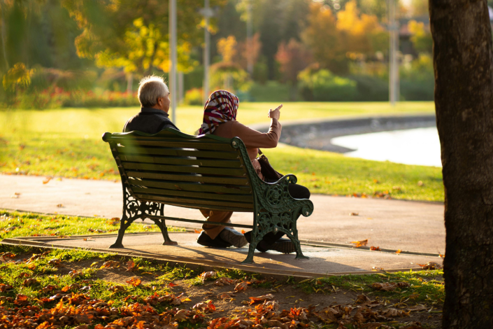 two people sitting on a bench in a park in front of a lake. one person is pointing forward.