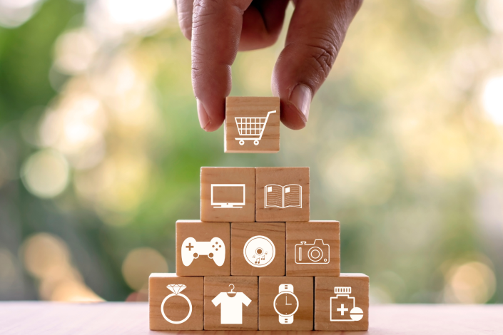 hand placing block on pile of blocks. each block has a symbol, including shopping cart, computer, book, gaming controller, CD, shirt, ring, camera, watch, and pill bottle