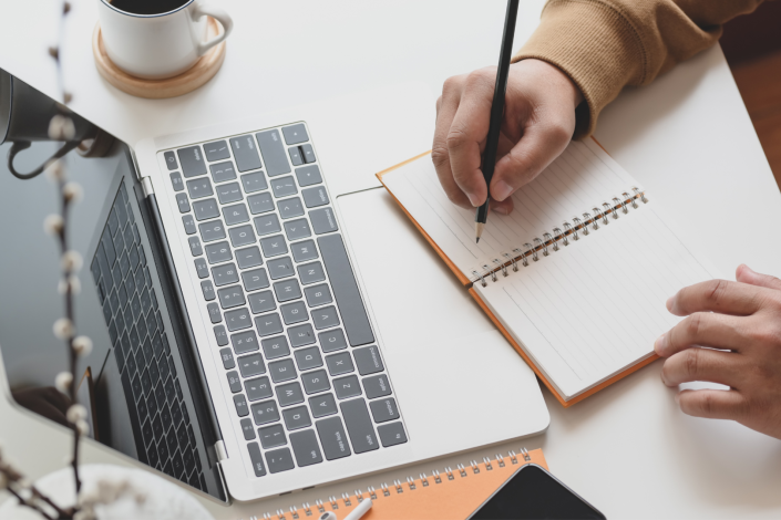 person holding a pencil above an open notebook. a laptop sits open in front of the notebook